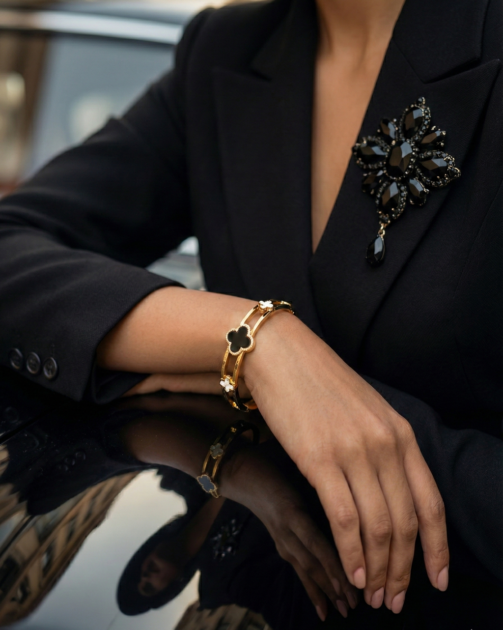 Person wearing a black blazer with a decorative brooch, gold bracelet, and gold ring.