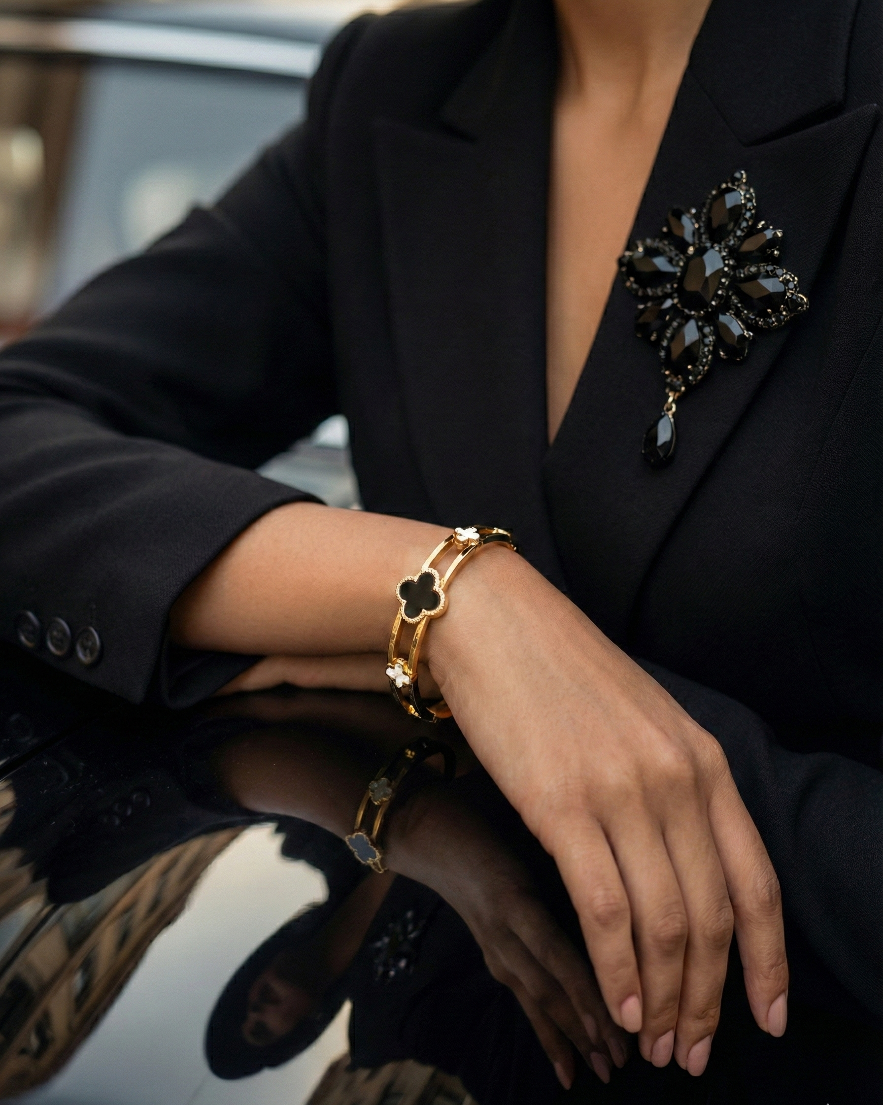 Person wearing a black blazer with a decorative brooch, gold bracelet, and gold ring.
